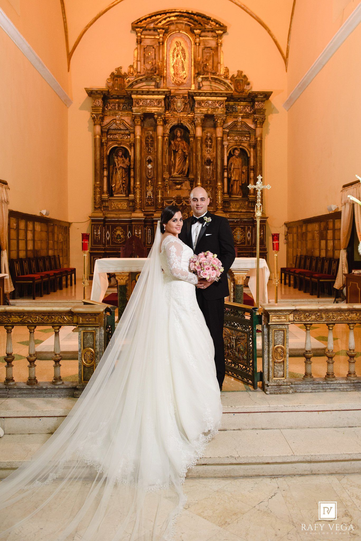 Bride in elegant gown with long veil and groom pose before ornate golden church altar.