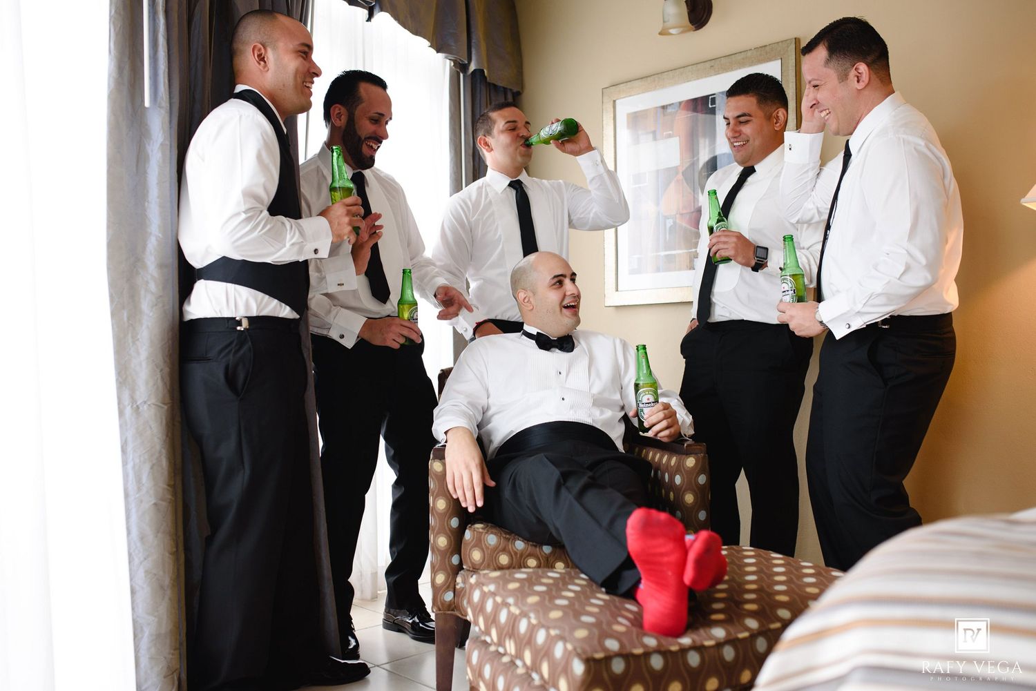 Groomsmen in black suits and ties celebrating with green beer bottles before a wedding ceremony indoors.