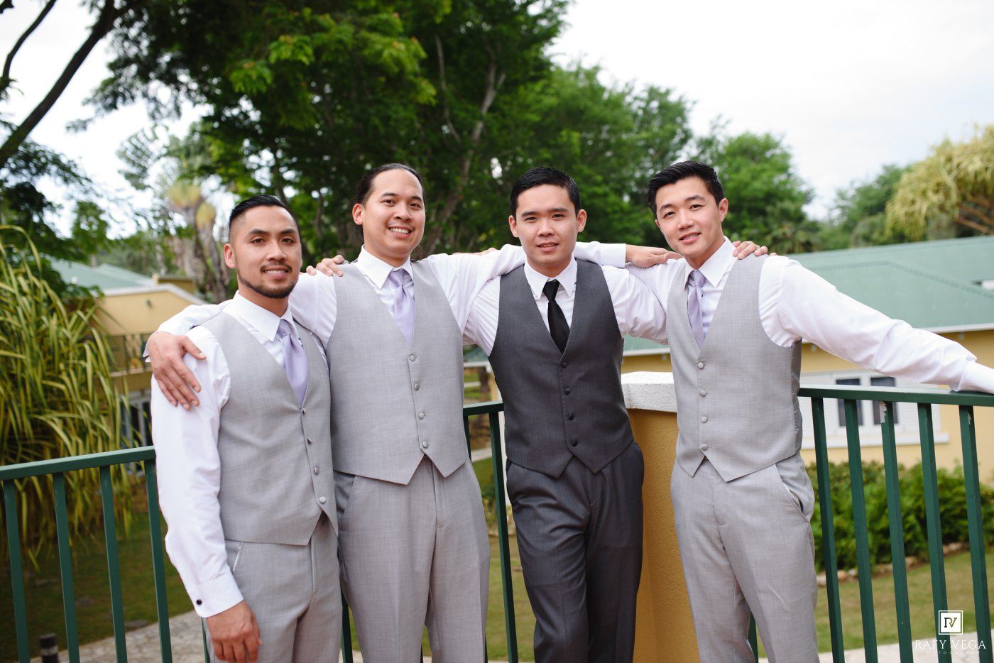 Four groomsmen in gray suits and vests pose together smiling on an outdoor balcony at a wedding.