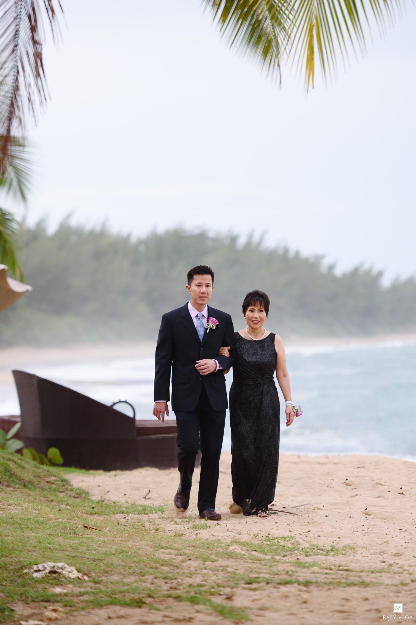 A groom in a dark suit and woman in black gown walk arm-in-arm along a tropical beach with palm trees.