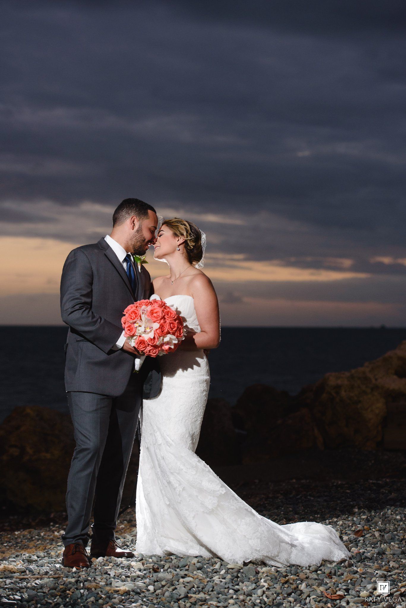 Bride and groom share a romantic kiss on a rocky beach at sunset, bride holding coral bouquet in white lace gown.