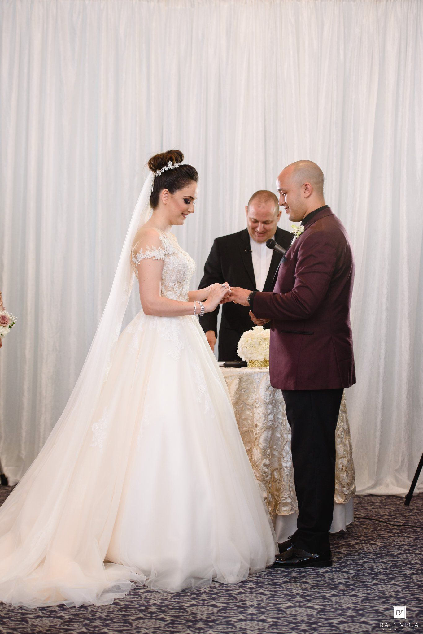 Bride and groom exchanging rings during wedding ceremony, officiant standing behind them, white drape backdrop.