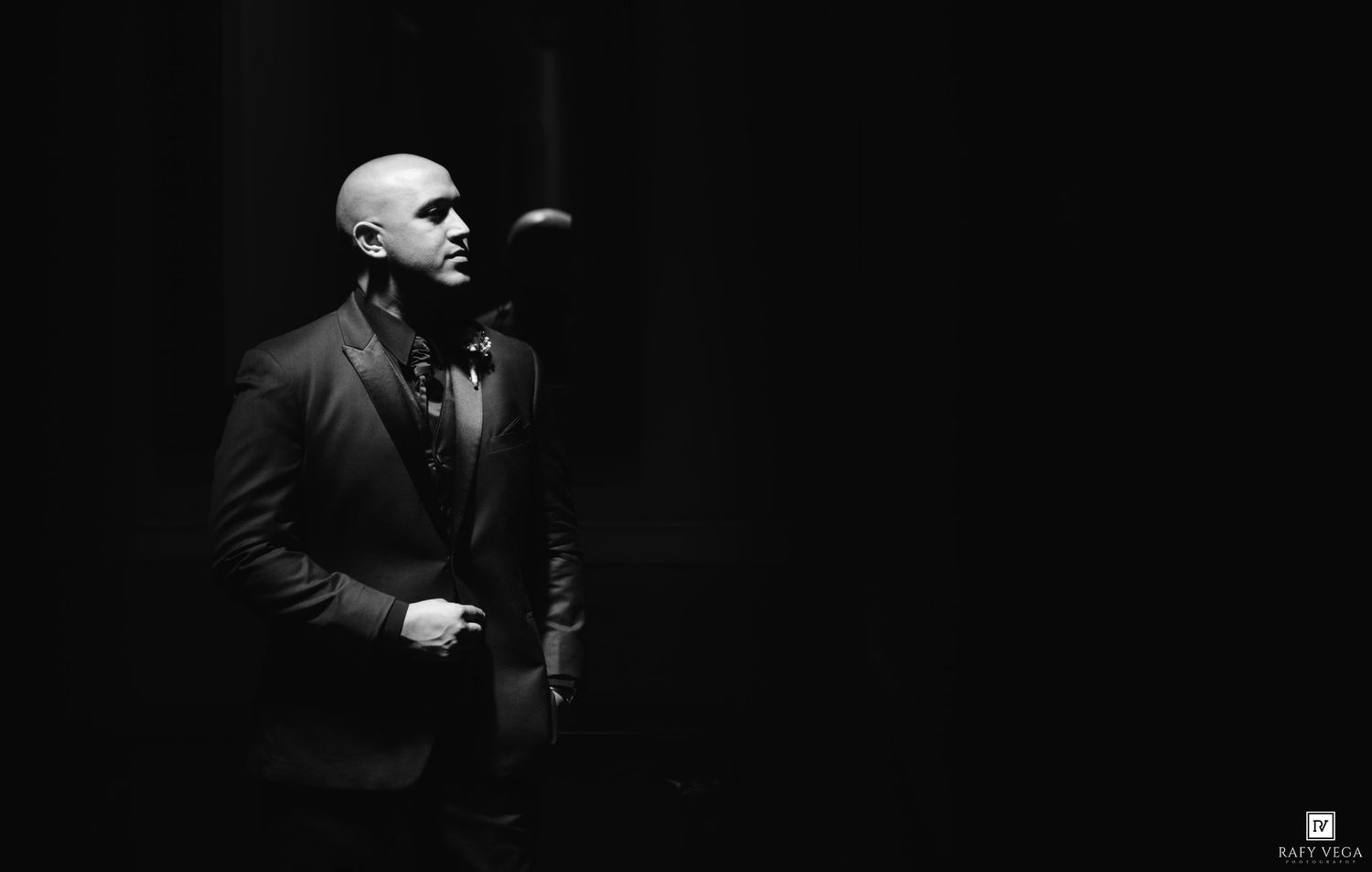 Black and white portrait of a bald man in a suit with bow tie standing in dramatic dark lighting.