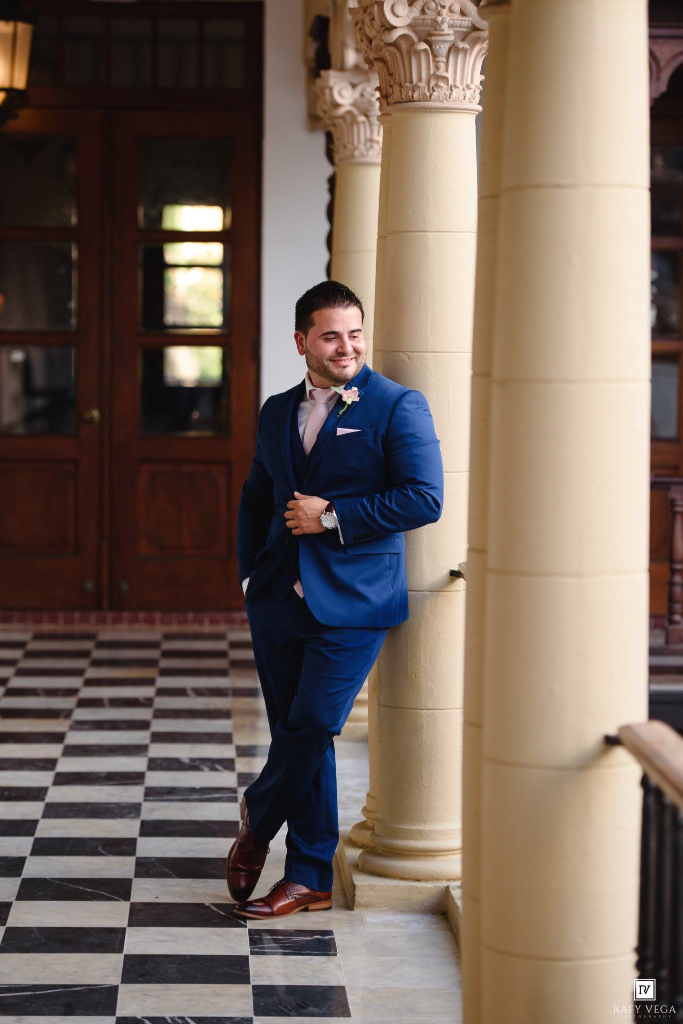 Dapper groom in navy blue suit leans against ornate column on checkered floor at elegant wedding venue.