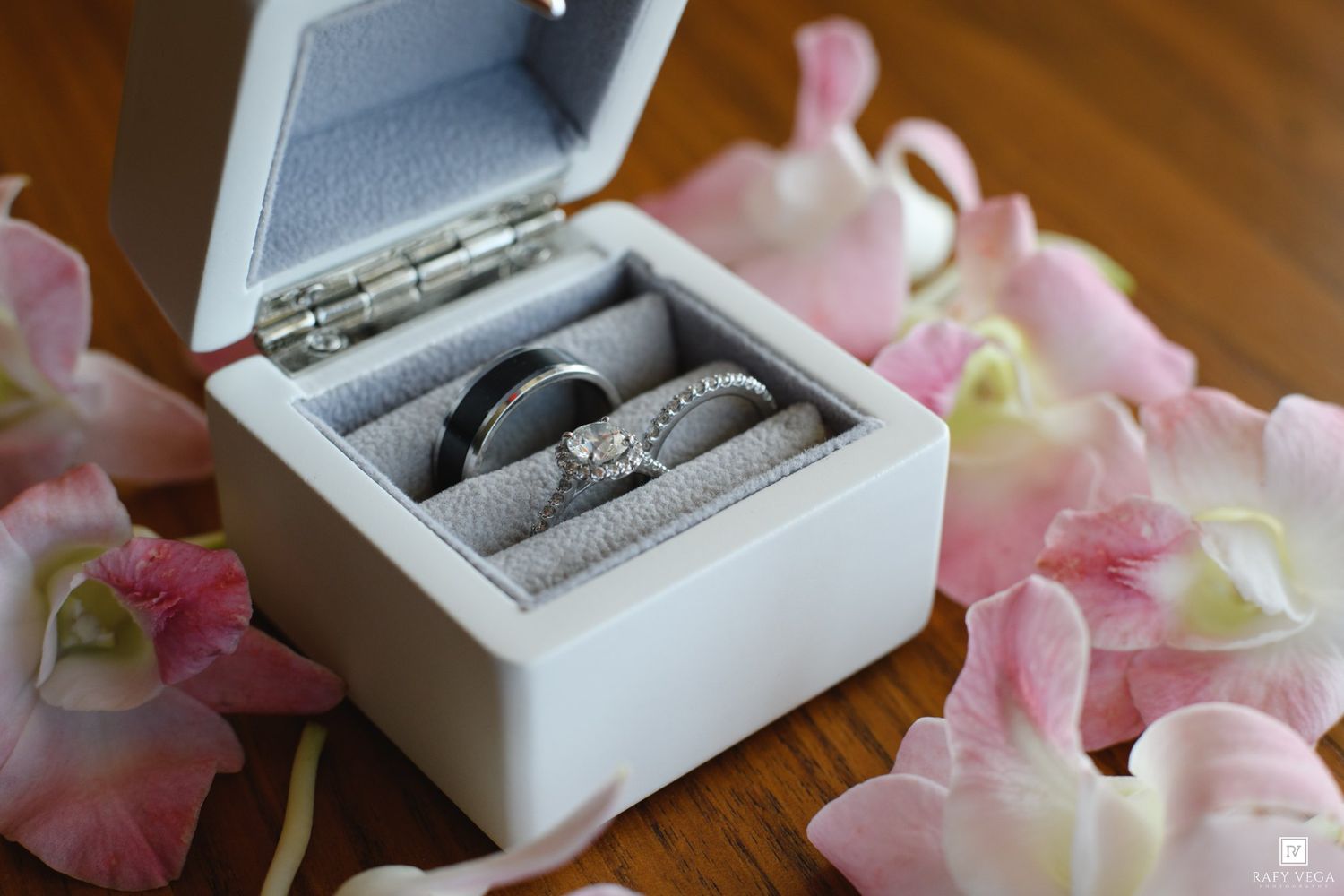 Two wedding bands and a diamond engagement ring displayed in a white velvet ring box surrounded by pink flower petals.