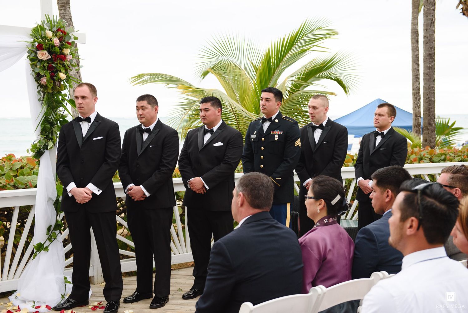 Groomsmen in tuxedos stand at an outdoor beach wedding ceremony with palm trees and floral arch.