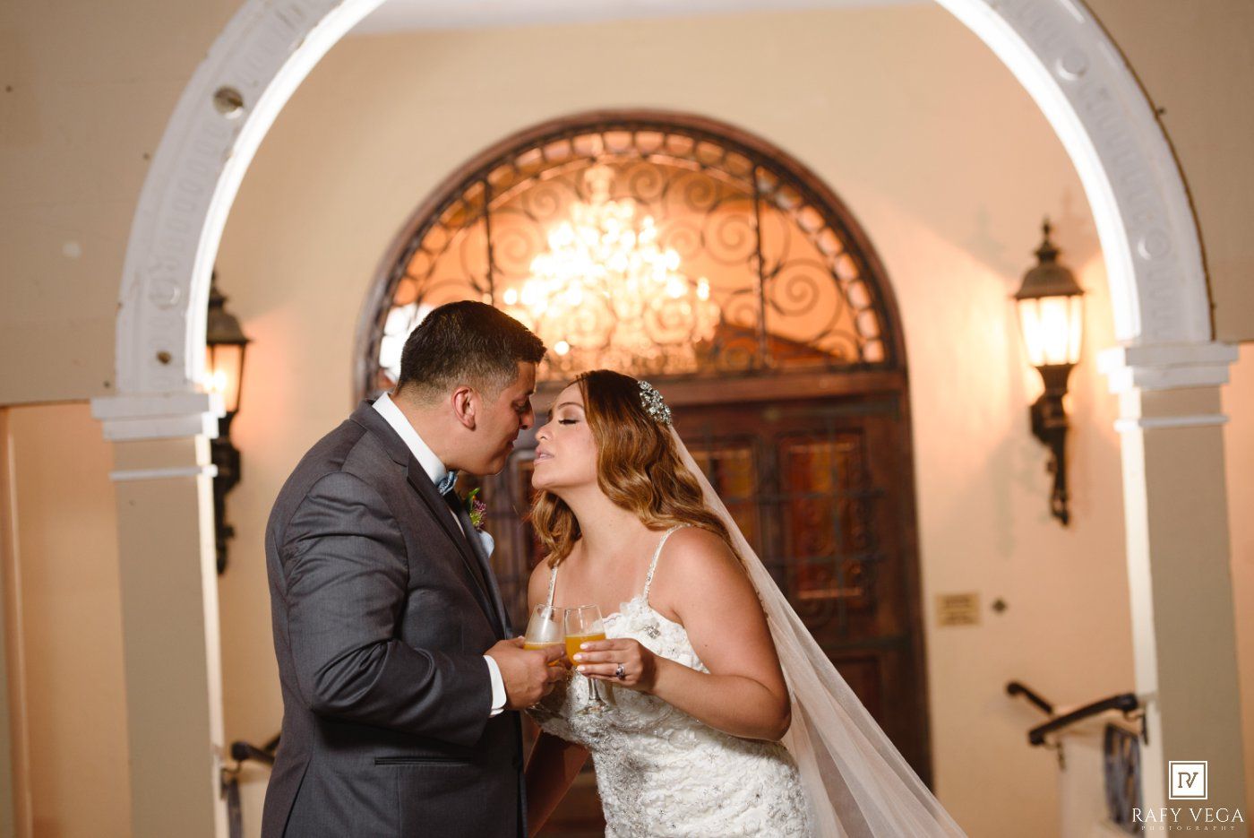 Bride and groom share a romantic moment under an elegant arched doorway with ornate stained glass window.