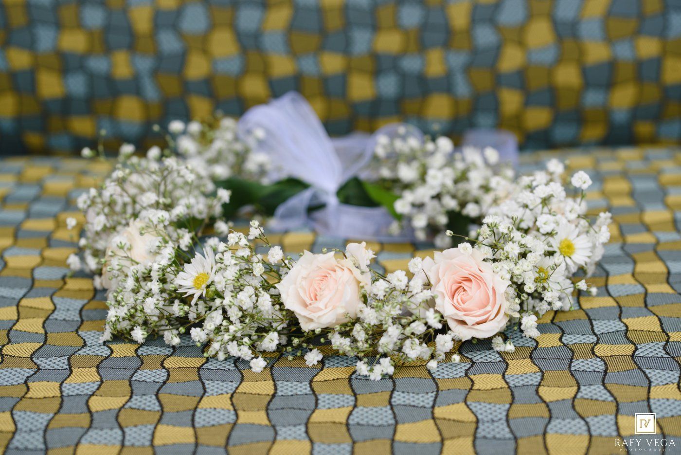 Delicate floral crown with pink roses and white baby's breath, adorned with a sheer ribbon, resting on a patterned fabric surface.
