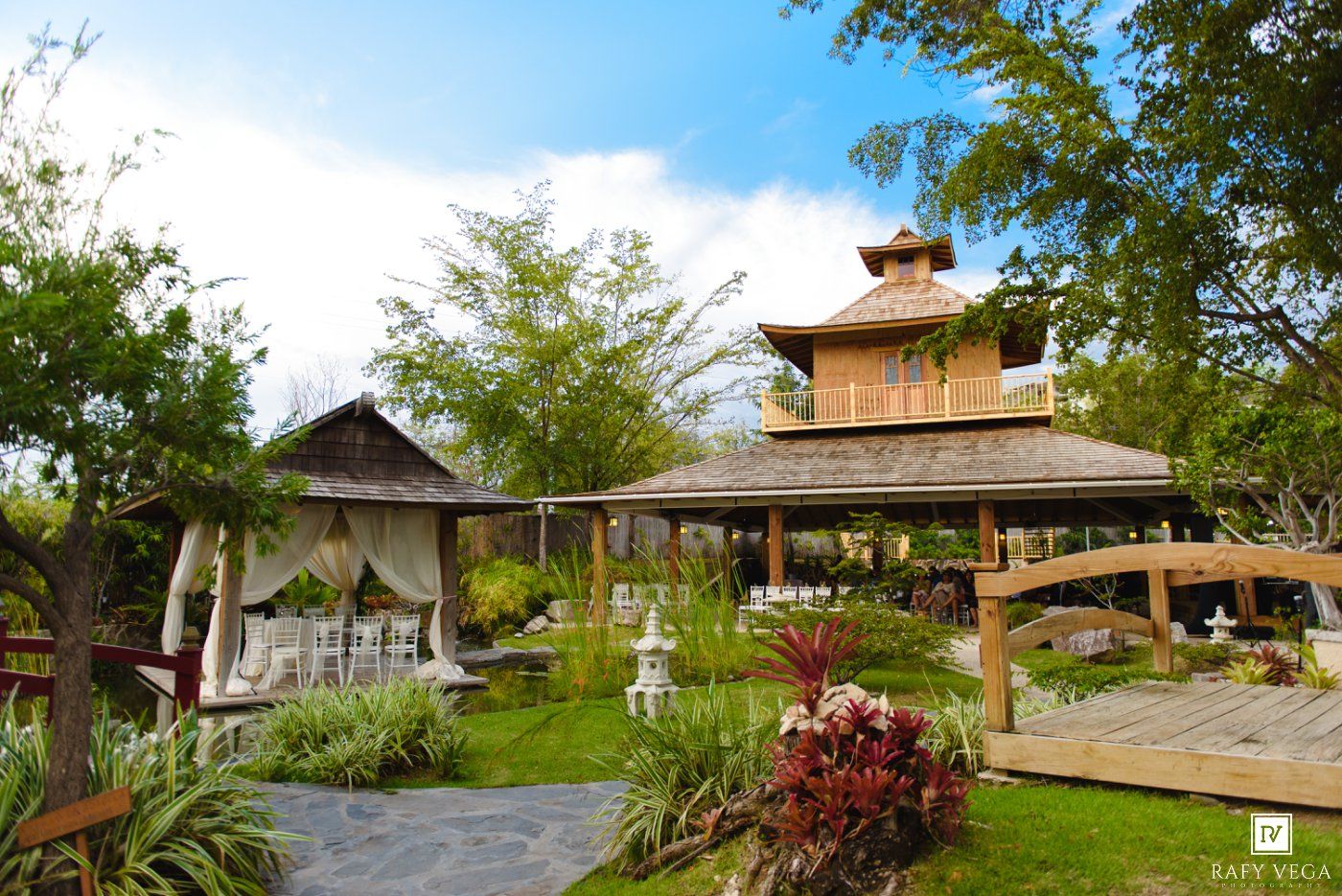 Elegant tropical wedding venue with gazebo, lush gardens, and wooden architecture under blue sky.
