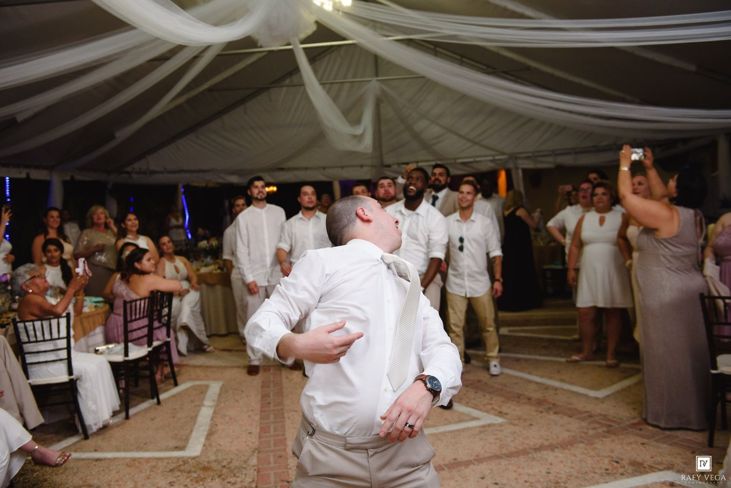 Two men embrace on a dance floor at an elegant wedding reception with white draping and guests watching.