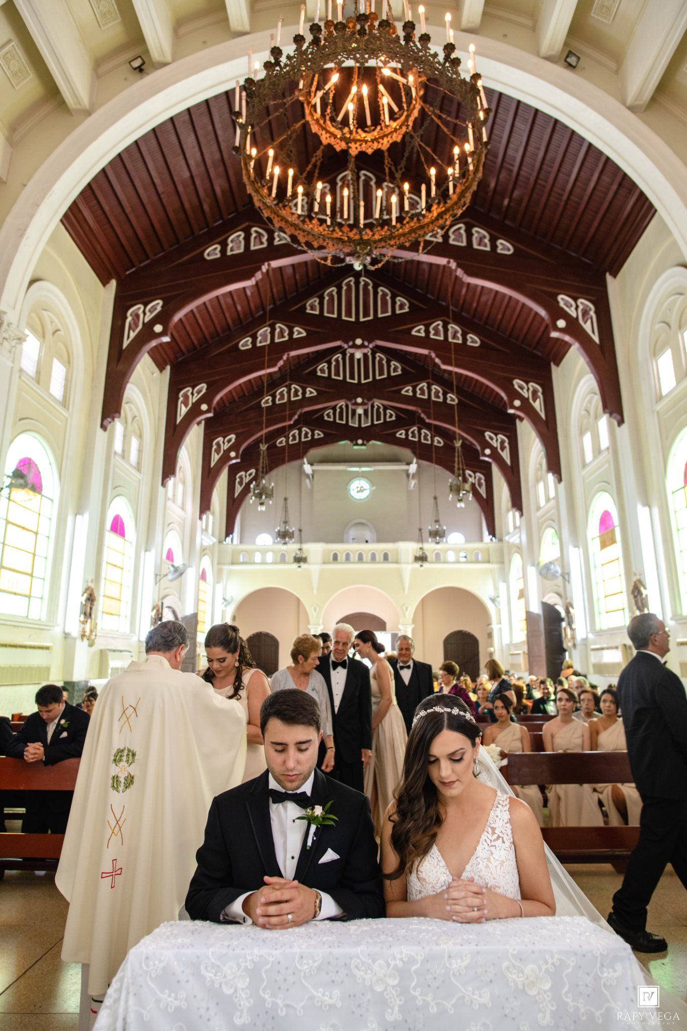 Bride and groom kneel at altar during elegant church wedding ceremony with ornate wooden ceiling and chandelier.