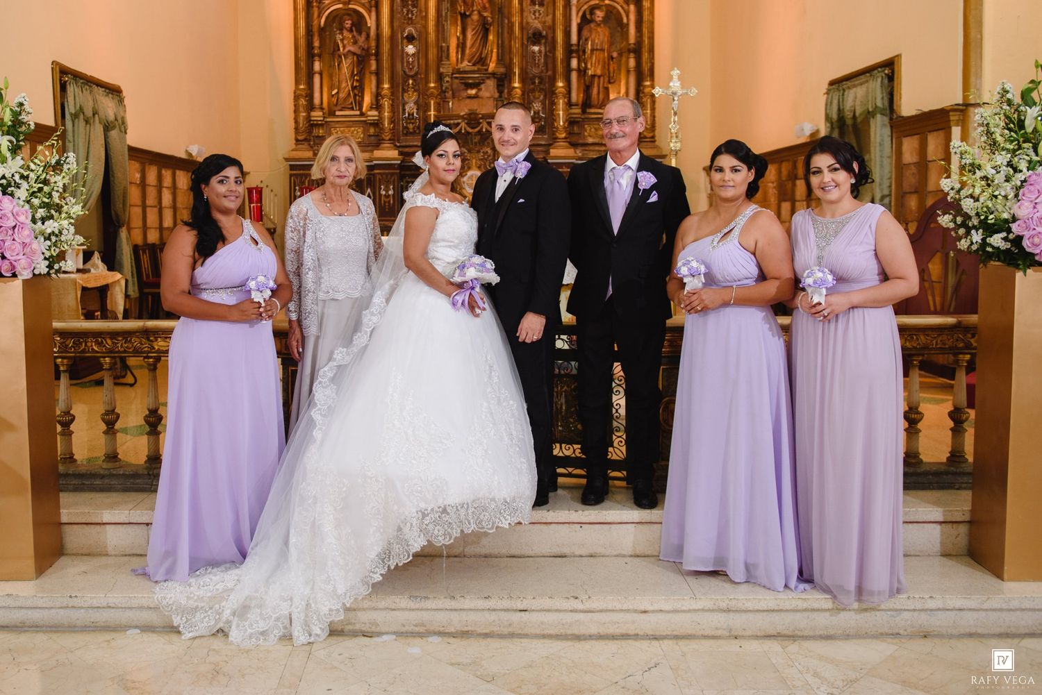 Wedding party poses in church with bride in white gown, groom in black suit, and four bridesmaids in lavender dresses.