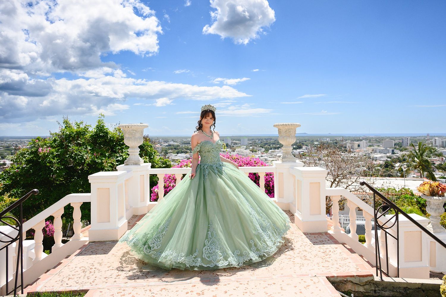 Young woman in a flowing mint green ball gown poses on elegant white terrace steps with panoramic city views.