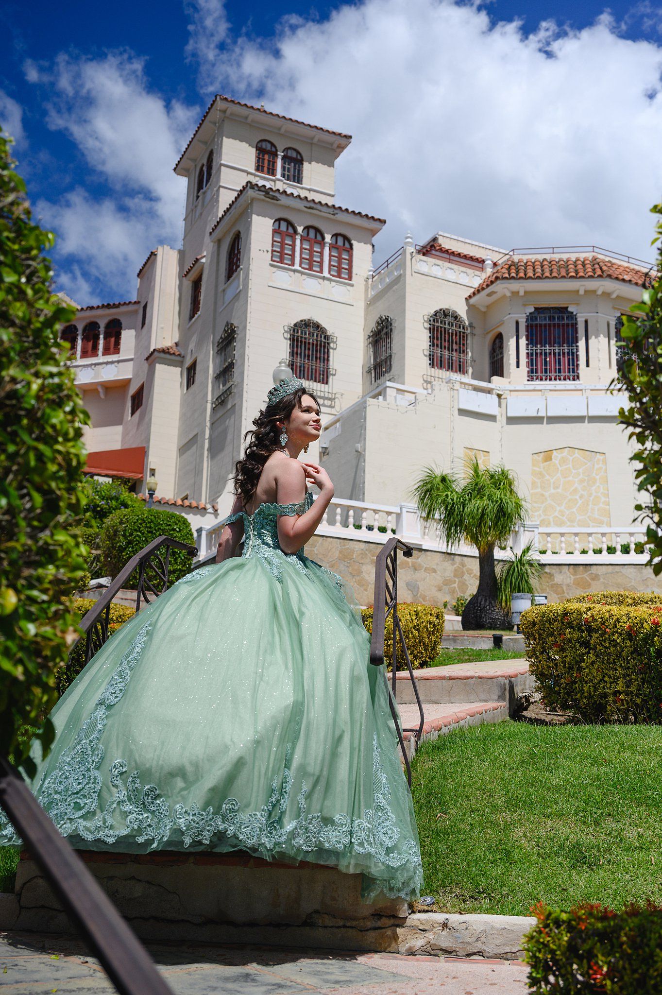 Young woman in a mint green quinceañera ball gown poses in a lush garden outside an elegant mansion.