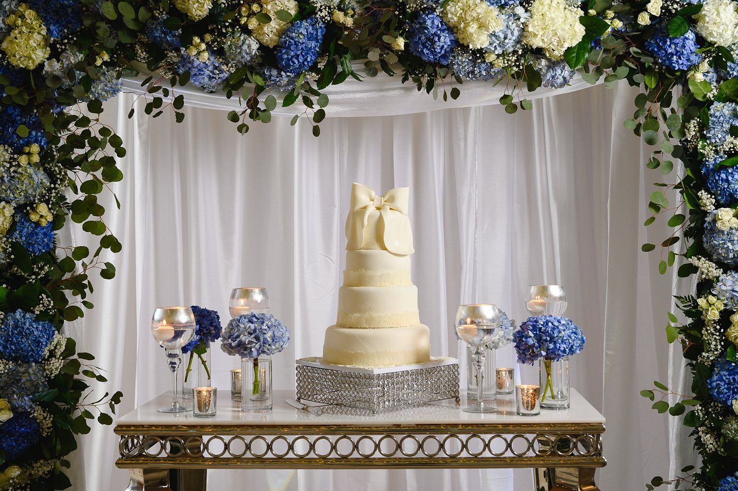 Elegant white tiered wedding cake with gold bow on crystal stand surrounded by blue hydrangeas under floral arch.