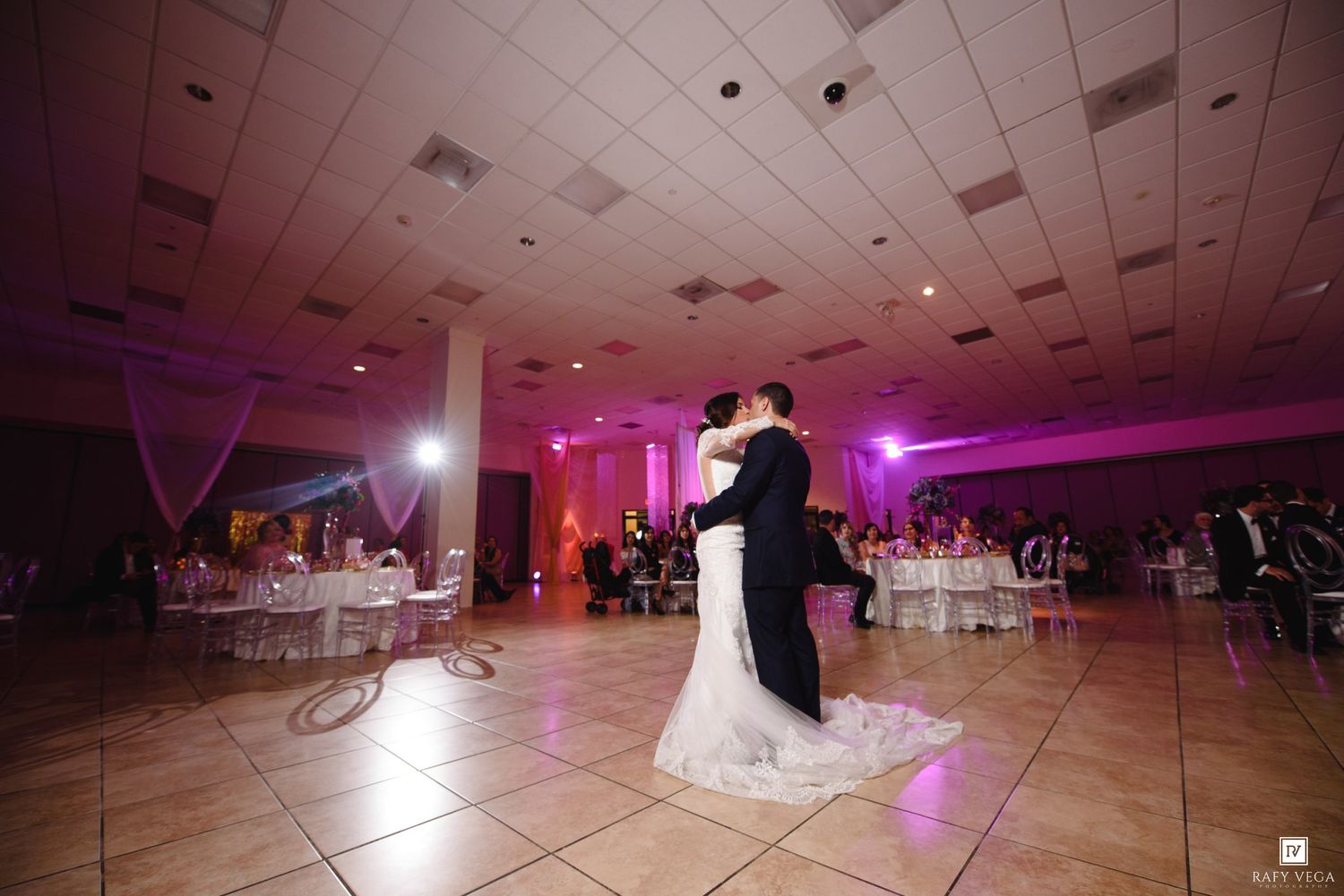 Bride and groom share their first dance on a lit dance floor at a romantic wedding reception with purple ambient lighting.