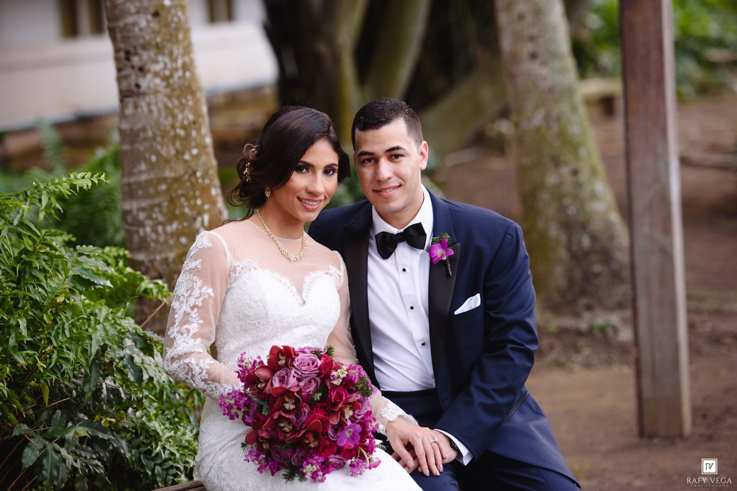 Bride and groom posing together outdoors, bride holding vibrant purple bouquet, groom in navy tuxedo.