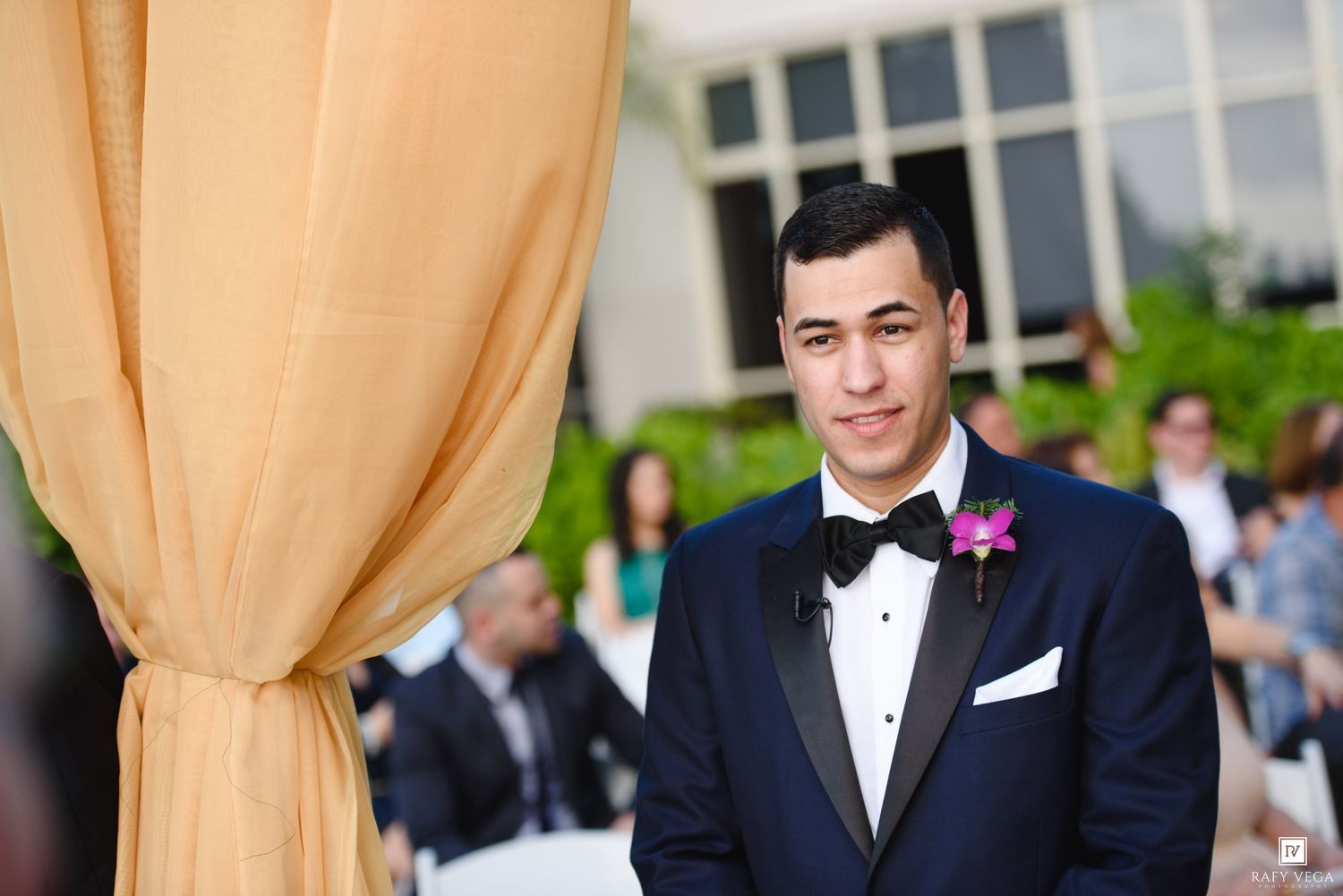 Handsome groom in navy tuxedo with bow tie and purple boutonniere stands at wedding altar.