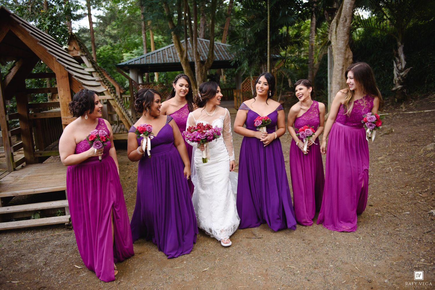 Bride in white gown with bridesmaids in purple and magenta dresses holding bouquets outdoors at a wedding.