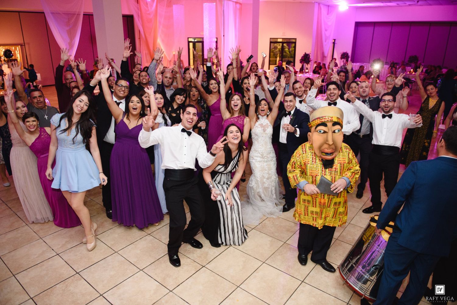 Wedding reception crowd cheering with bride and groom on dance floor, guests in purple bridesmaid dresses celebrating.