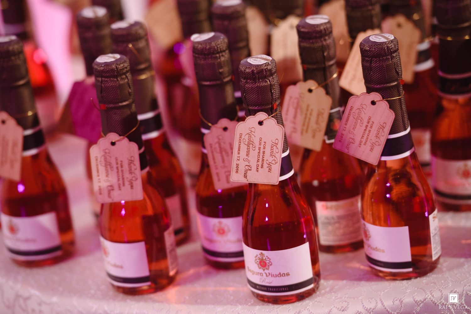 Small rose wine bottles with gift tags displayed as wedding favors on a decorated table.