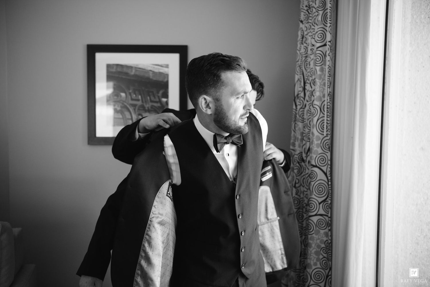 Groom getting helped into his suit jacket by groomsman before wedding ceremony in black and white photo.
