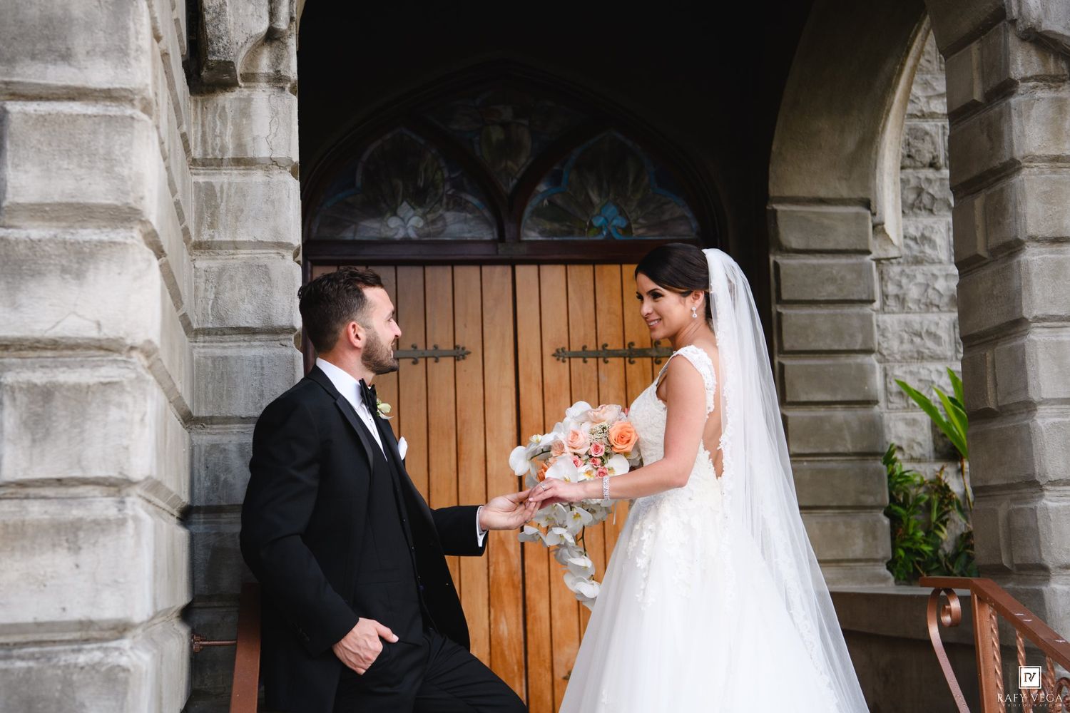 Bride and groom share a romantic moment outside a stone church, bride holding a peach bouquet in a backless white gown.
