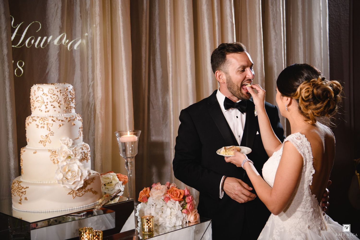 Bride feeding groom wedding cake slice at luxurious reception with ornate white and gold tiered cake.