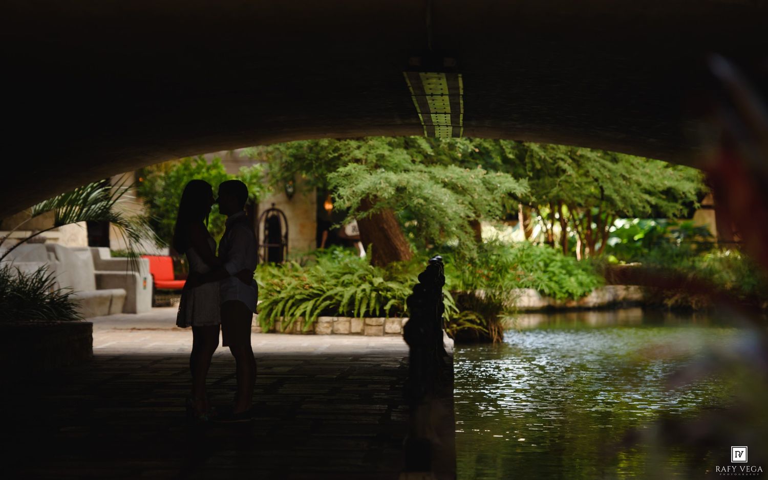 Silhouettes of two people standing near a serene pond under a bridge arch surrounded by lush tropical greenery.