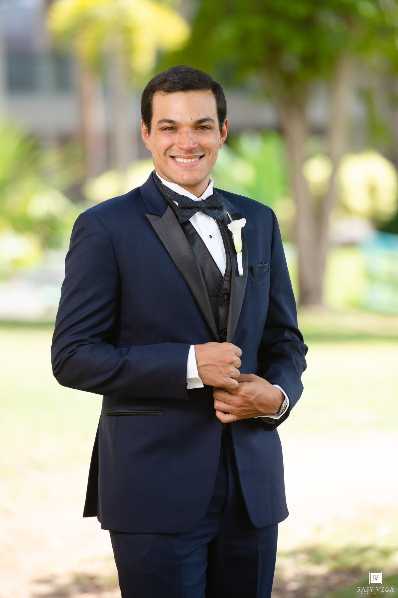Smiling groom in navy blue tuxedo with black lapels and bow tie, wearing a white boutonniere outdoors.
