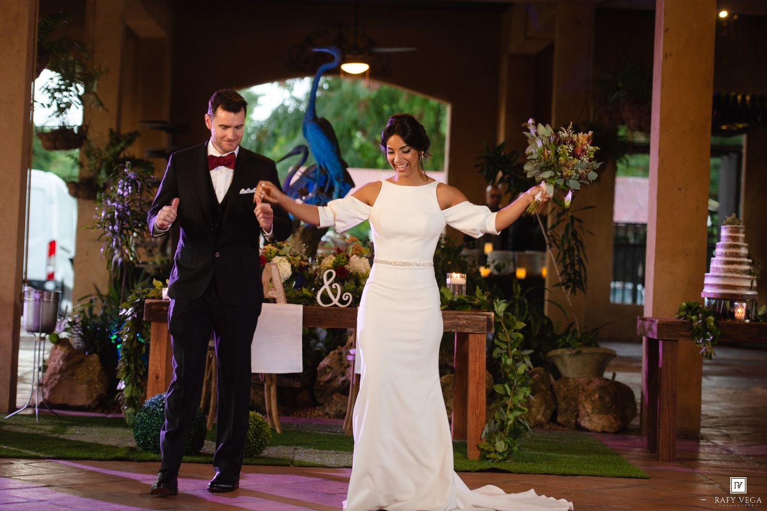 Bride in white gown and groom in black tuxedo hold hands while making grand entrance at elegant outdoor wedding reception.