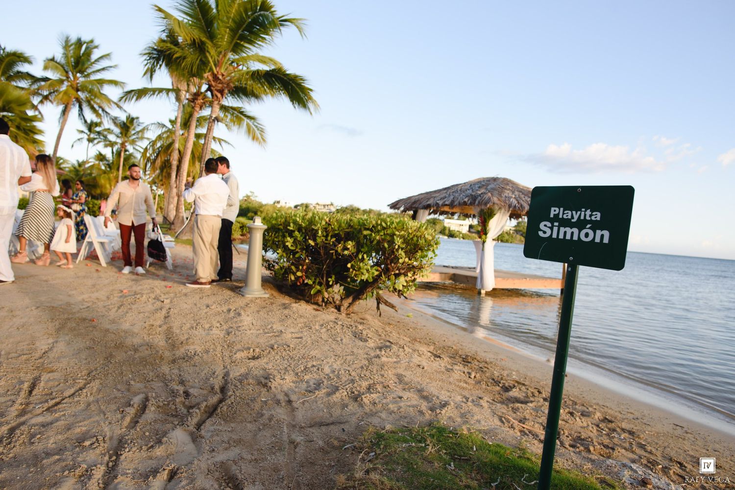 Sign for Playita Simon beach with palm trees and wedding guests gathered along the tropical shoreline.