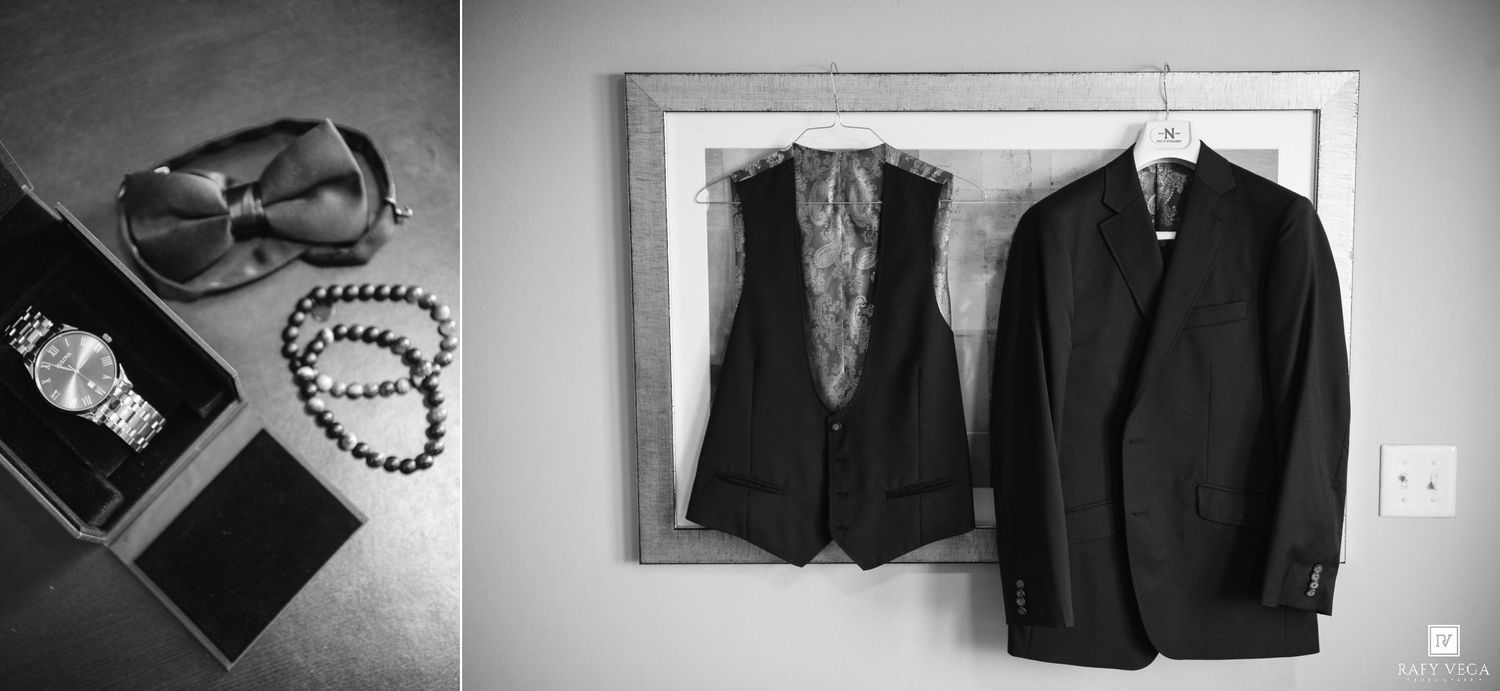 Black and white photo of groom accessories including sunglasses, watch, bracelet, and suit jacket with vest hanging on wall.