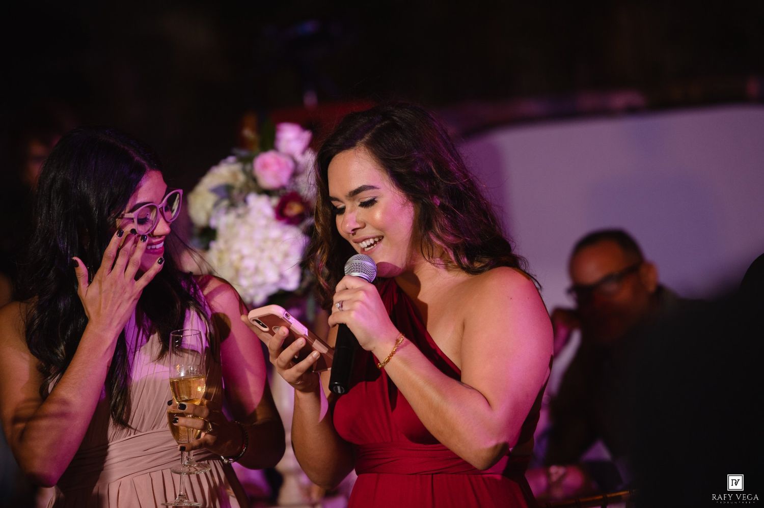 Woman in red dress speaks into microphone at elegant event while friend reacts with surprise beside her.