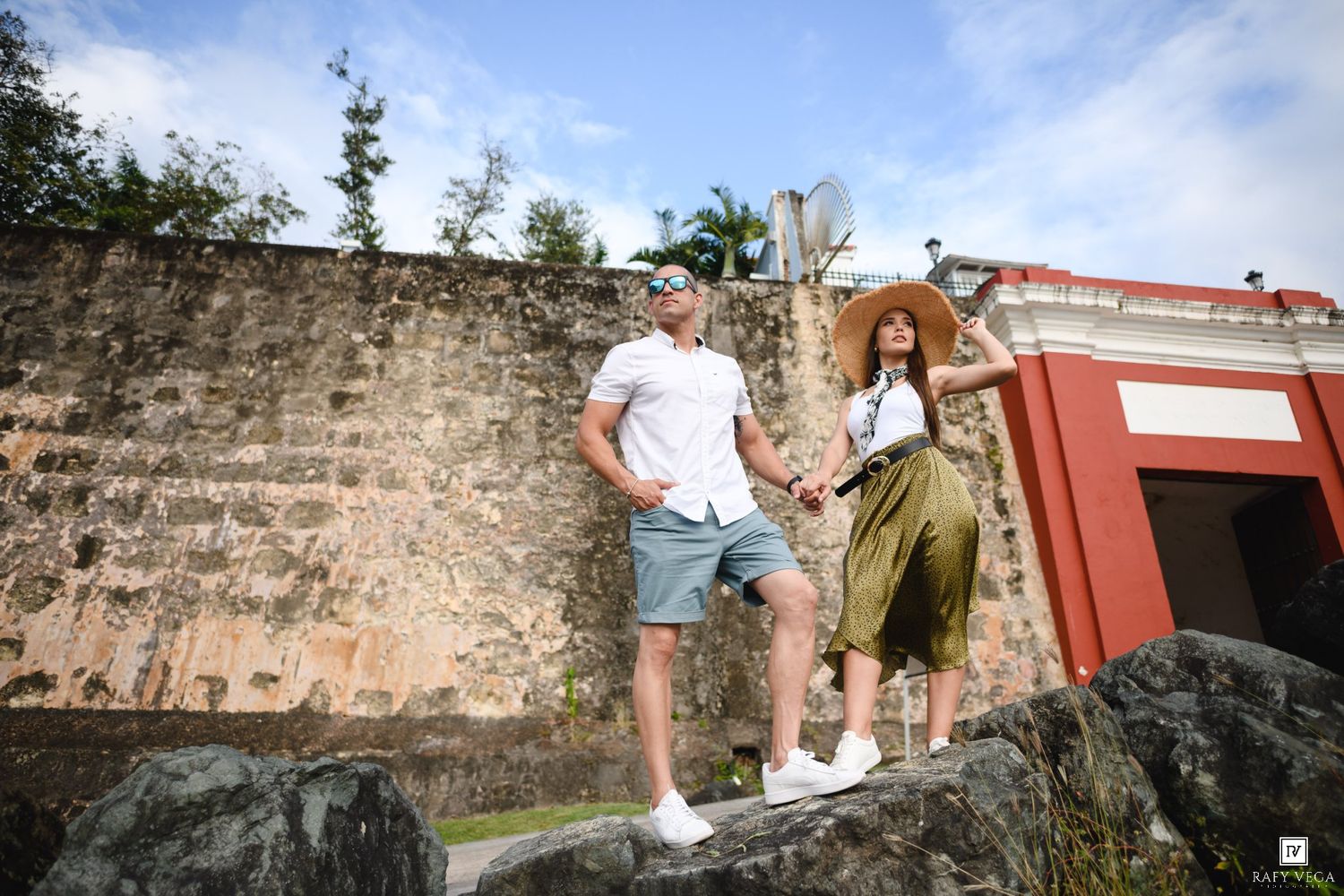 Couple posing on rocks near historic stone wall and red building outdoors.