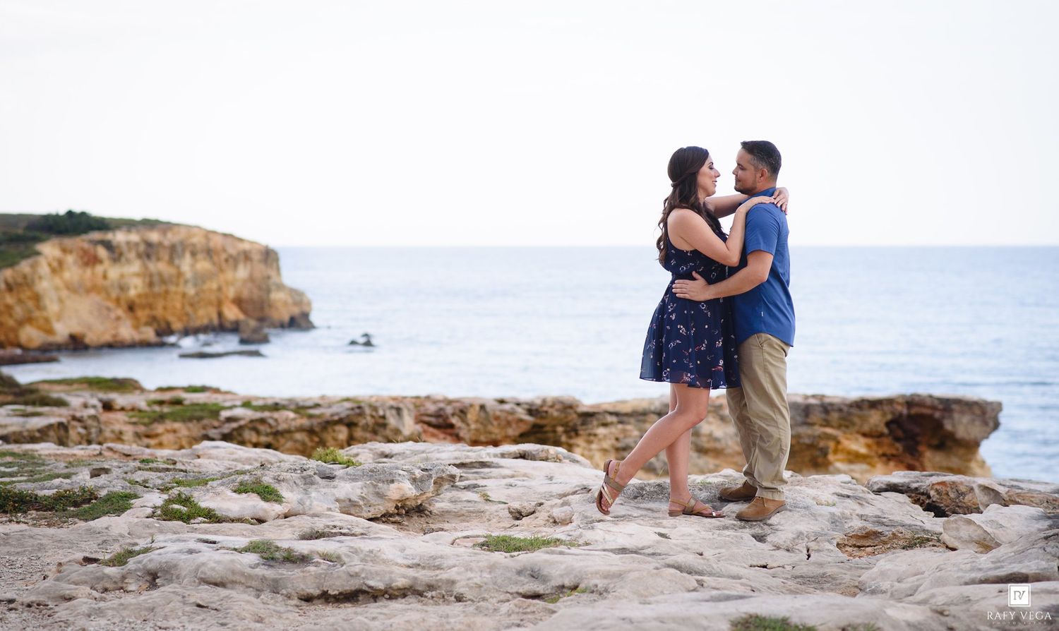 Couple dancing and embracing on rocky coastal cliffs with turquoise ocean views during a romantic engagement session.