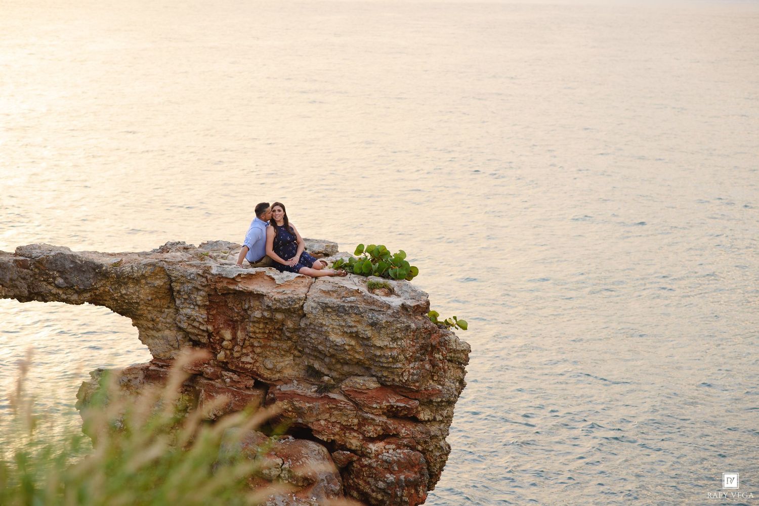 Couple sitting together on a rocky ocean cliff at sunset, sharing a romantic moment with calm sea waters below.