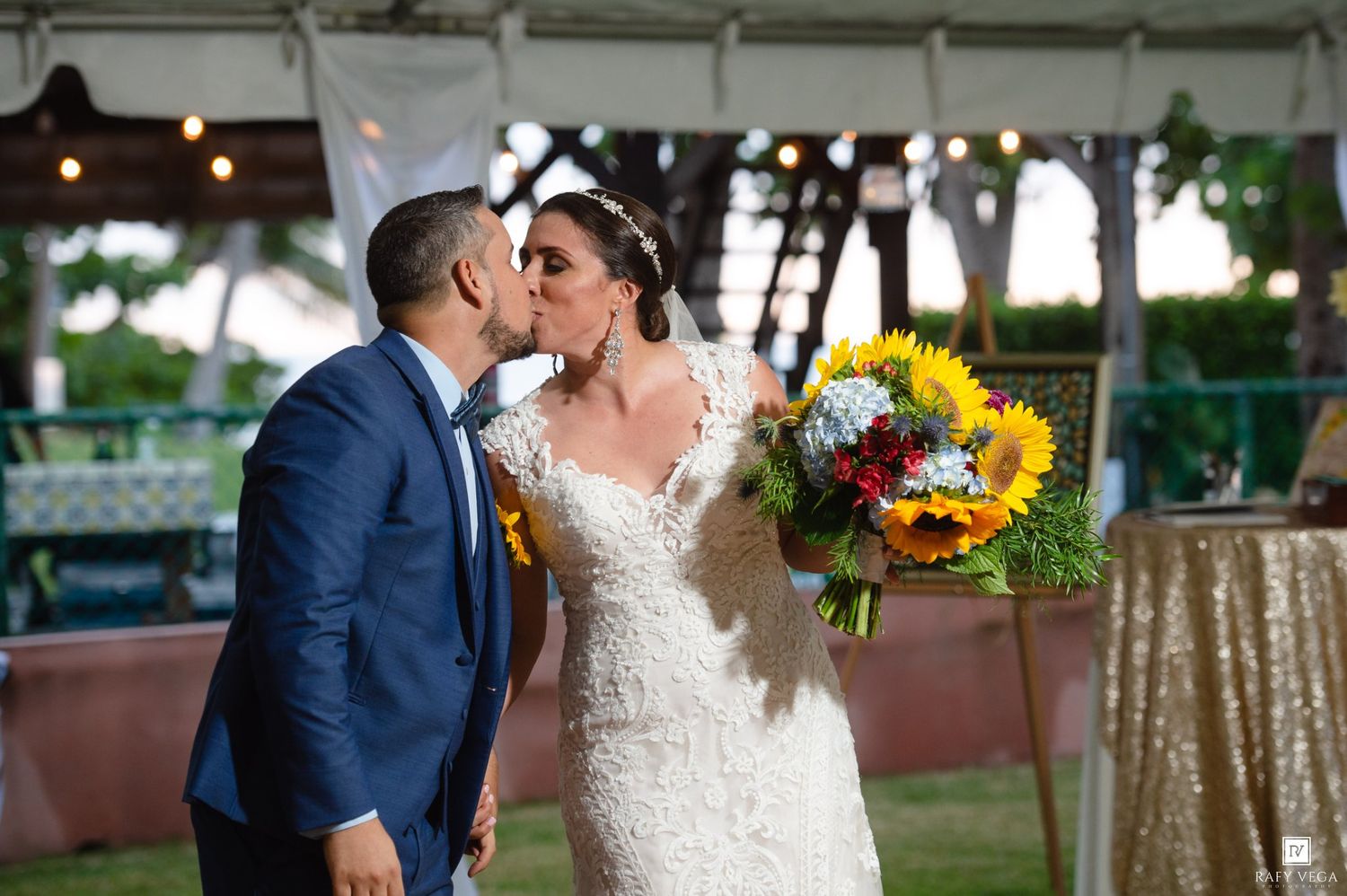 Bride and groom share a romantic kiss outdoors at wedding ceremony, bride holding vibrant sunflower bouquet.