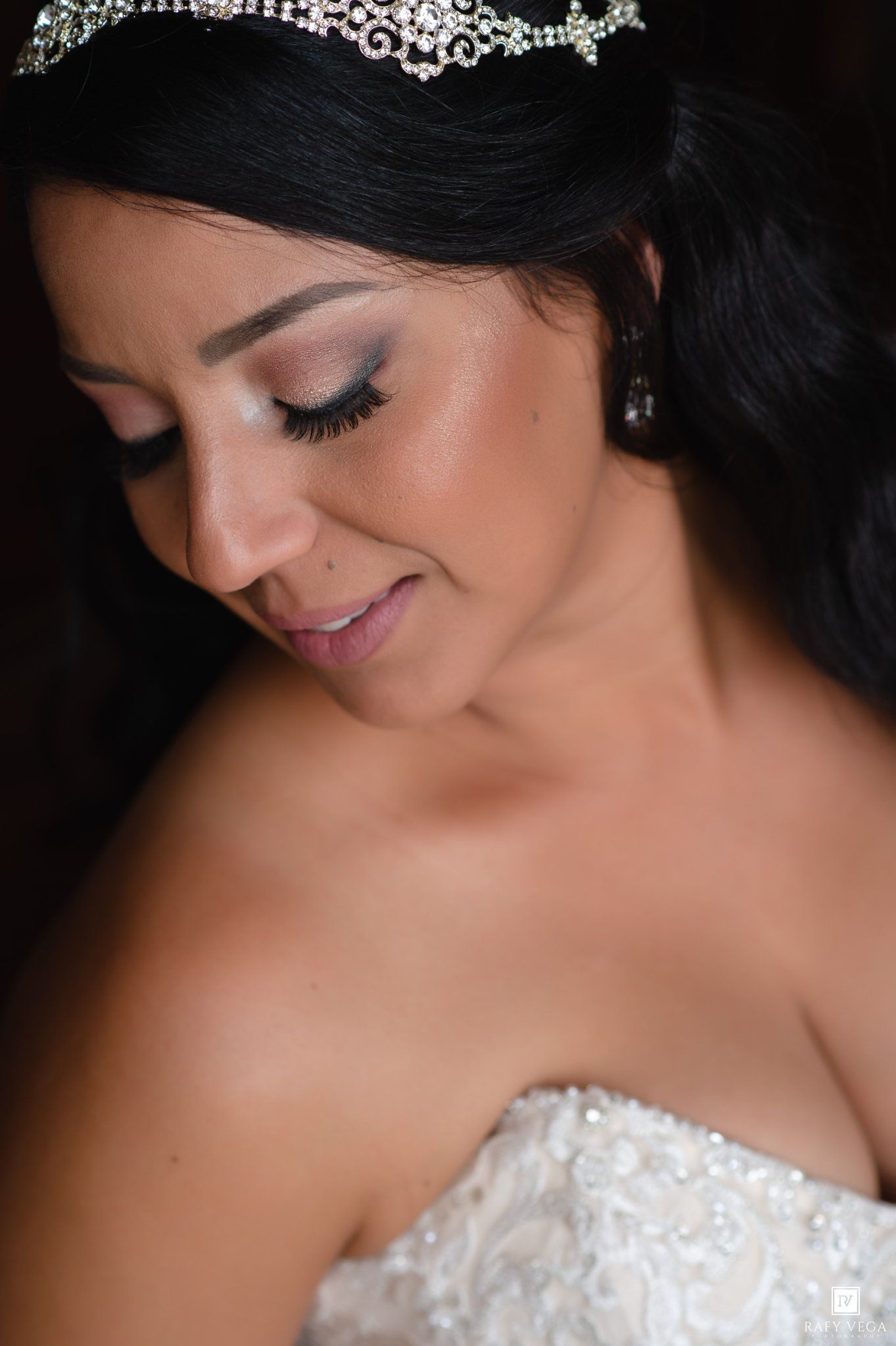 Bride in a strapless lace gown wearing a crystal tiara and drop earrings, smiling softly with elegant bridal makeup.