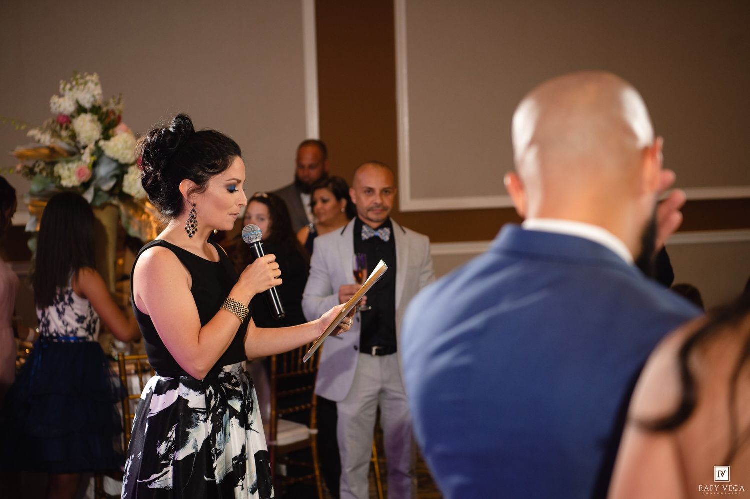 Woman in black dress reads from paper into microphone at elegant wedding reception event.