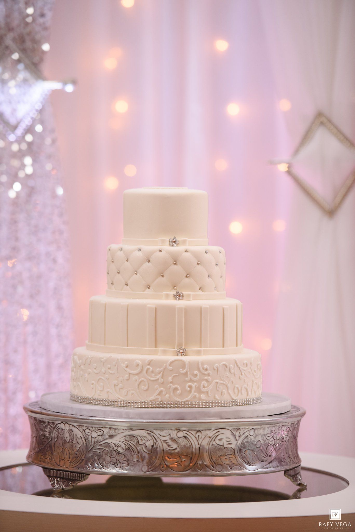Elegant four-tier white wedding cake with quilted and scroll details on a silver stand against a pink backdrop.