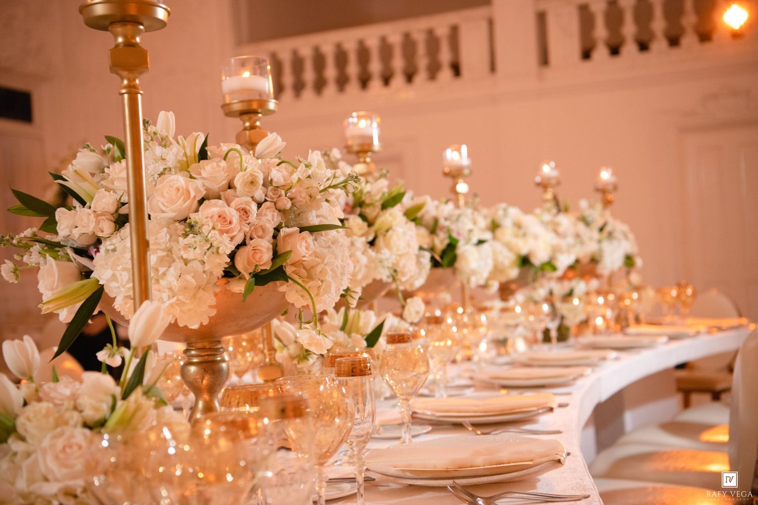 Elegant wedding reception table with white floral centerpieces, gold candelabras, and glowing candlelight in a grand ballroom.