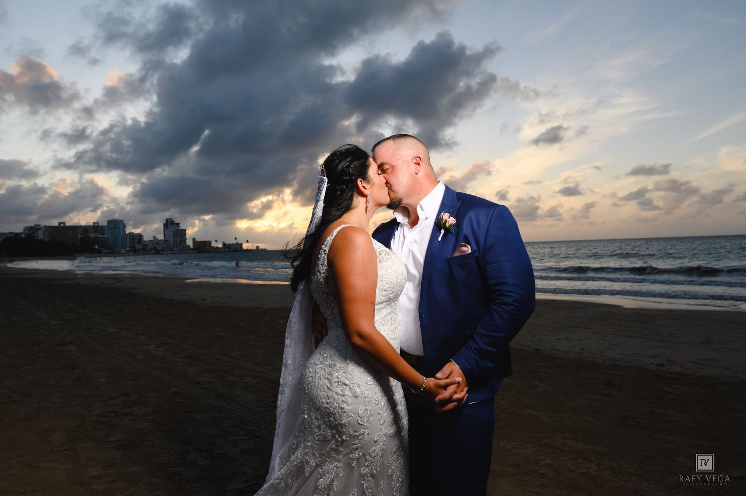 Bride and groom share a romantic beach kiss at sunset with city skyline and dramatic cloudy sky backdrop.