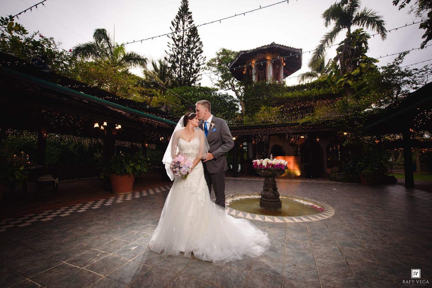Bride and groom share a romantic kiss in an elegant outdoor courtyard with lush greenery and a fountain at dusk.