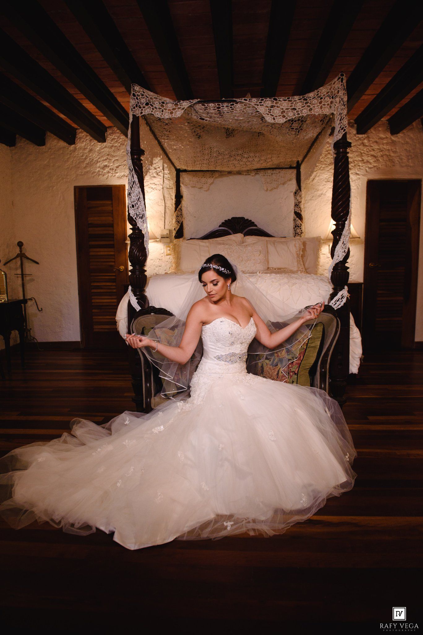 Elegant bride in strapless ballgown sits before canopy bed in rustic stone room on wedding day.