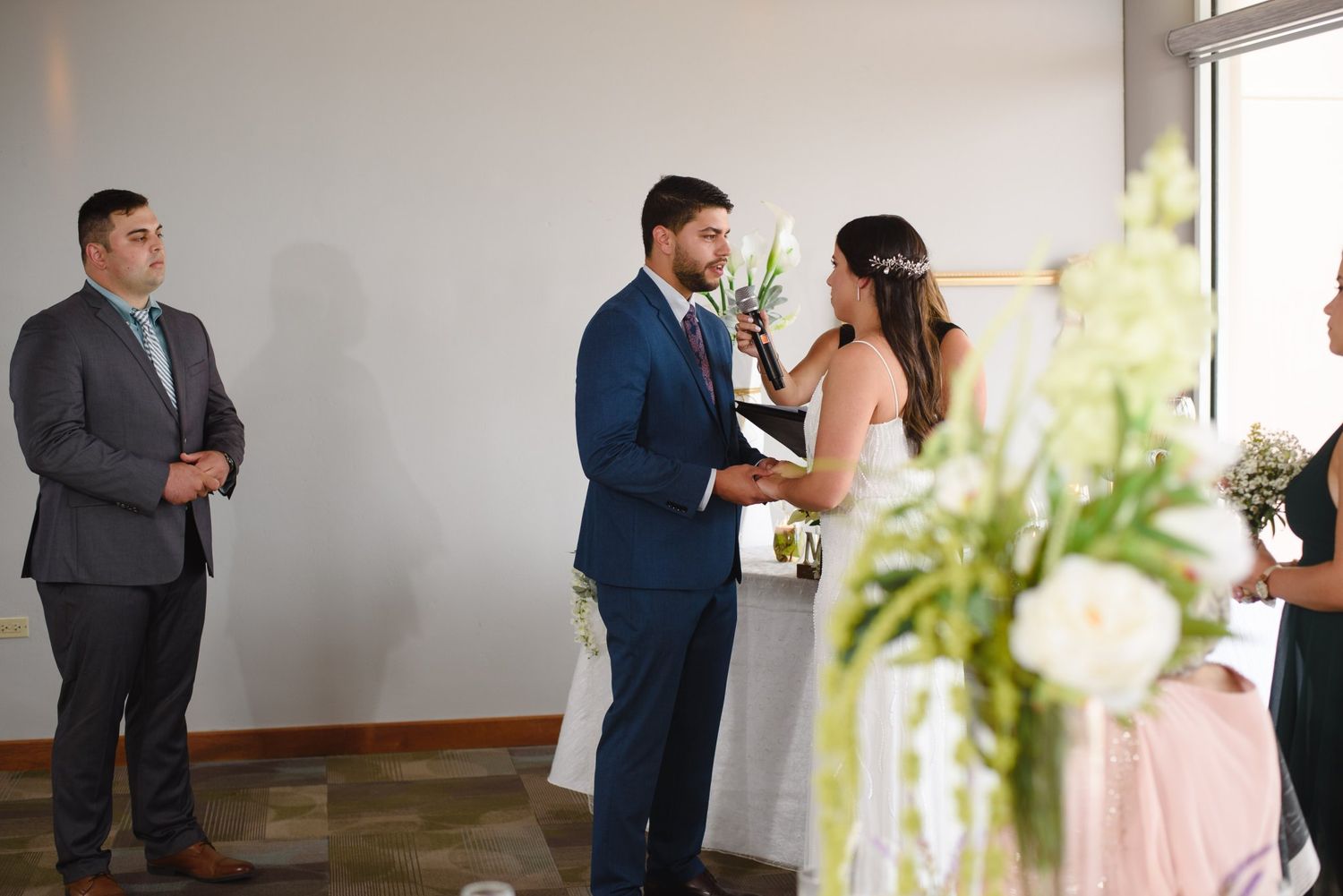Bride and groom exchange vows at indoor wedding ceremony with officiant and wedding party in background.