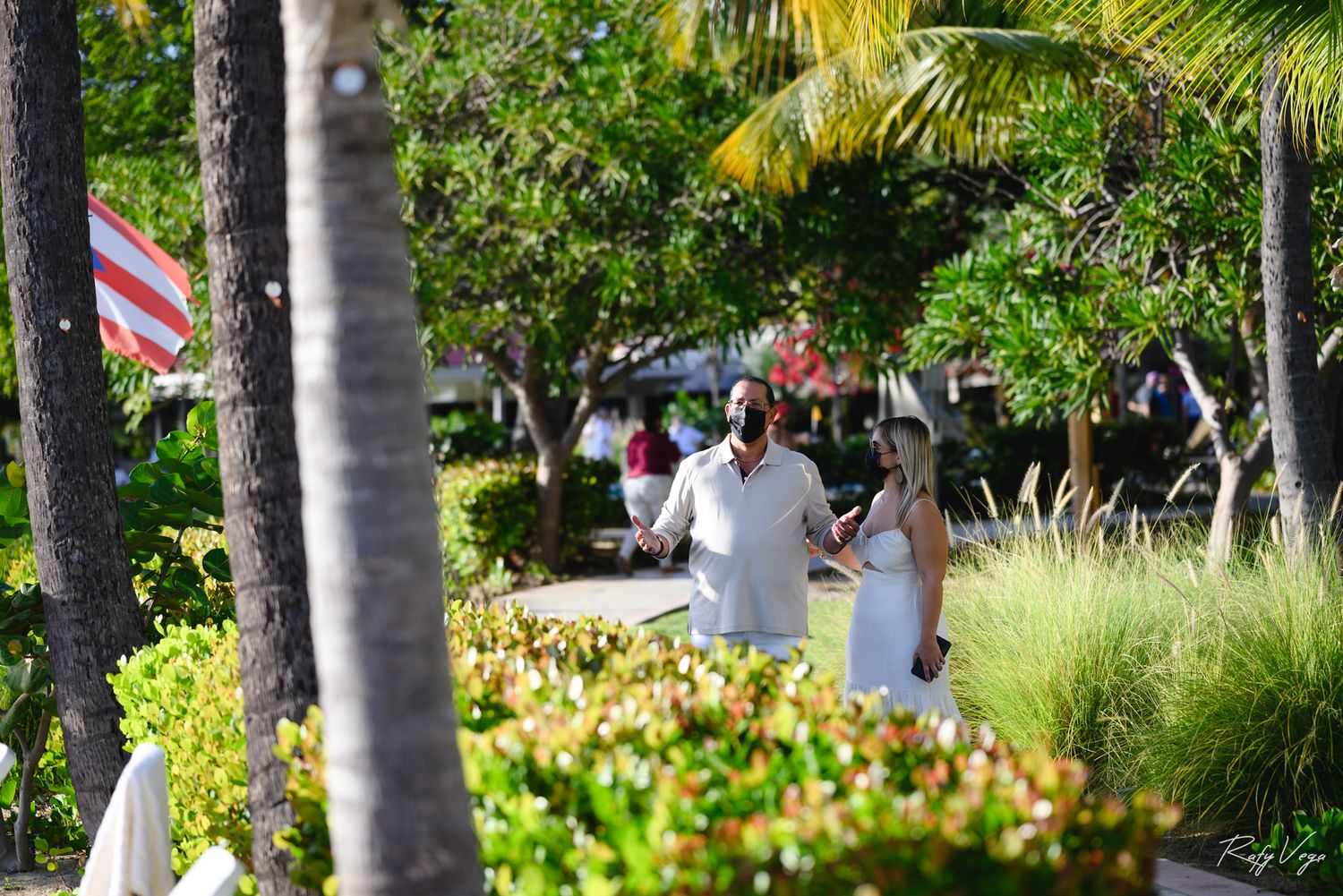 Couple walking through lush tropical garden path with palm trees and colorful plants in bright sunlight.