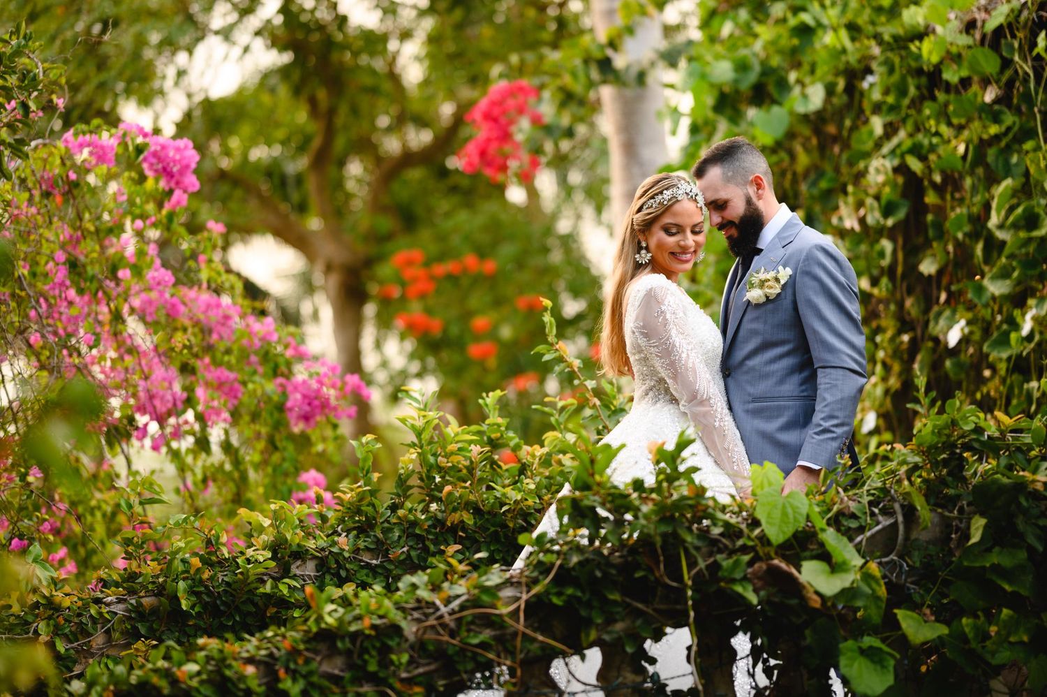 Bride and groom posing romantically among lush greenery and colorful bougainvillea flowers in a garden setting.