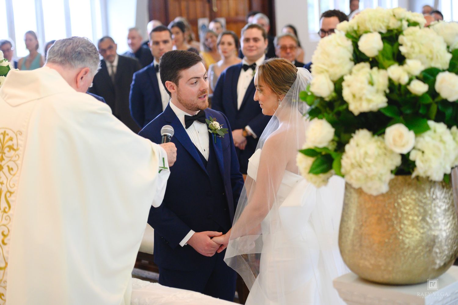 Couple exchanging wedding vows at church altar, groom in navy suit, bride in white gown, white floral arrangement nearby.