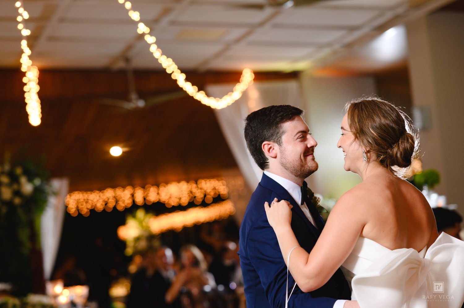 Bride and groom share their first dance, smiling at each other under warm string lights at their wedding reception.